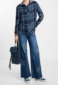 Persona che indossa una camicia a quadri blu, jeans a gamba larga ricamati con motivi floreali, sneakers blu e tiene uno zaino blu navy per il manico.