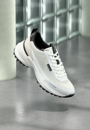 Witte en zwarte Calvin Klein sneaker met zijvakken van mesh en een dikke zool, zwevend boven een grijze vloer nabij een glasblokmuur.