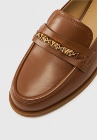 Καφέ δερμάτινα μοκασίνια με διακοσμητική αλυσίδα σε χρυσό χρώμα και λεπτομέρειες με λογότυπο στο μπροστινό μέρος. Στρογγυλεμένη μύτη και ραμμένο περίγραμμα.