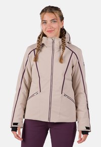 Vrouw met gevlochten haar, gekleed in een beige jas met capuchon en paarse accenten, voorzien van ritszakken, gecombineerd met donkerpaarse broek.