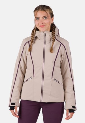 Mujer con cabello trenzado que lleva una chaqueta con capucha color beige con ribetes morados y bolsillos con cremallera, combinada con pantalones morados oscuros.