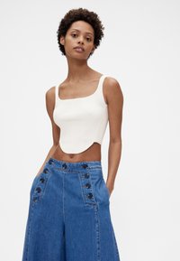 Hvid ribbet crop top med firkantet halsudskæring, kombineret med højtaljede blå denim bukser med vide ben og knapdetaljer i siden.
