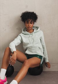 Um hoodie verde claro com um bolso canguru, combinado com calções verdes escuros com riscas brancas, sentado numa bola de exercício preta.