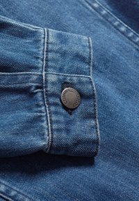 Tissu en denim bleu foncé avec un gros plan sur une manchette, mettant en avant un bouton en métal noir embossé avec "CORNWALL SEAL." Détails de couture visibles.