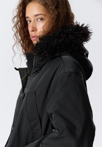 Parka neagră cu glugă căptușită cu blană sintetică, având o închidere prin buton și un buzunar lateral. Textură netedă și croială ajustată.