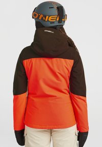 Veste de neige orange et noire avec capuche ajustable, texture douce et logo dans le dos. Associée à un pantalon de neige beige et des gants noirs.