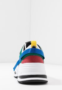 Zapatilla multicolor con suela blanca, que presenta acentos en verde, rojo, negro y azul. Incluye una correa amarilla en el talón y materiales texturizados.