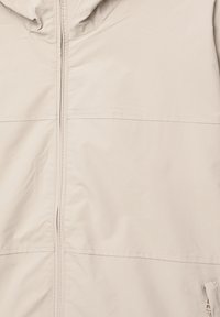 Veste beige clair avec une fermeture éclair à l'avant, une capuche et des coutures sans couture. Fabriquée en tissu lisse et léger, présentant des lignes épurées et un design minimal.
