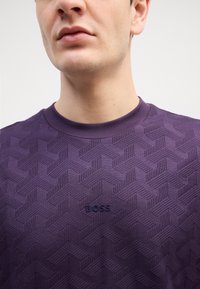 T-shirt a girocollo di colore viola con texture, caratterizzato da un motivo geometrico a zigzag e un piccolo logo ricamato "BOSS" dello stesso colore.