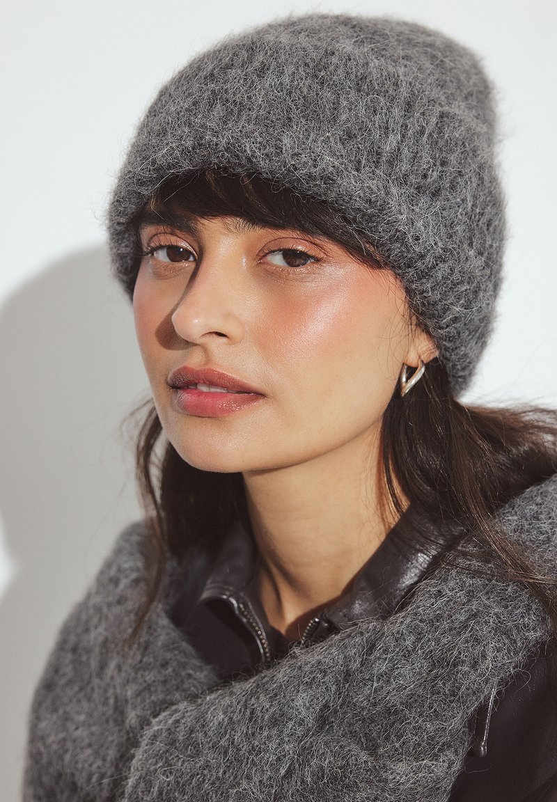 Beanie di lana grigia con una finitura morbida e testurizzata; abbinato a una sciarpa grigia coordinata. Il modello indossa una giacca di pelle nera. Sfondo chiaro.
