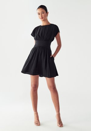 Donna in piedi che indossa un vestito nero corto con vita arricciata e maniche a cappotto, tacchi nude con cinghie e orecchini a cerchio dorati su uno sfondo bianco.