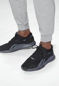 Füße, die schwarze und graue Sportschuhe mit schwarzen Knöchelsocken und hellgrauen, strukturierten Jogginghosen auf einer weißen Oberfläche tragen.