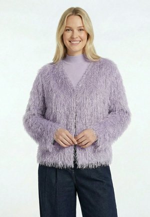 Femme souriante portant un cardigan duveteux lavande sur un col roulé violet clair et un pantalon foncé, debout devant un fond clair uni.