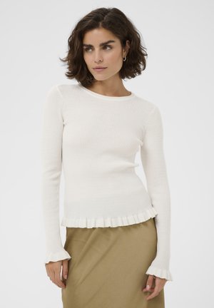 Žena s krátkymi kučeravými vlasmi, ktorá má na sebe biele rebrované tričko s dlhým rukávom so záhybmi na manžetách a spodnom okraji, doplnené béžovou sukňou.