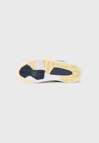 Semelle de basket avec motifs beige, marine et blanc et un logo circulaire près du talon sur fond gris clair.