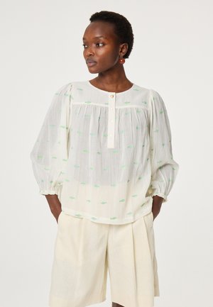 Woman wearing a white blouse with green fish patterns, puffed sleeves, pearl buttons, and cream-colored shorts, hands in pockets, looking left.