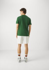 Zöld, rövid ujjú pamut keverék póló, fehér sorttal párosítva. A modell mintás zoknit és sportcipőt visel, hátulról nézve.