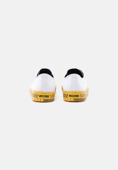 Ζευγάρι λευκά slip-on sneakers, ορατά από πίσω, με κίτρινο σχέδιο μετρητικής ταινίας και λογότυπο MOSCHINO στις λαστιχένιες σόλες.