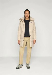 Jauns cilvēks ar pinumiem matos, ģērbies bežā piepūšamā kapuce, melnā krekla, khaki biksēs un pelēkos sporta apavos, stāv uz balta fona.