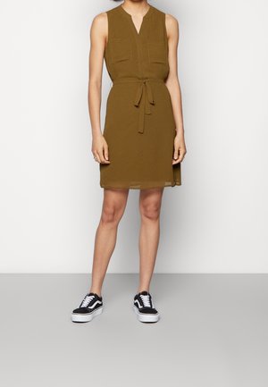 Ærmeløs olivengrøn kjole med forlommer og taljebånd, parret med sort-hvide platform sneakers på en stående person.