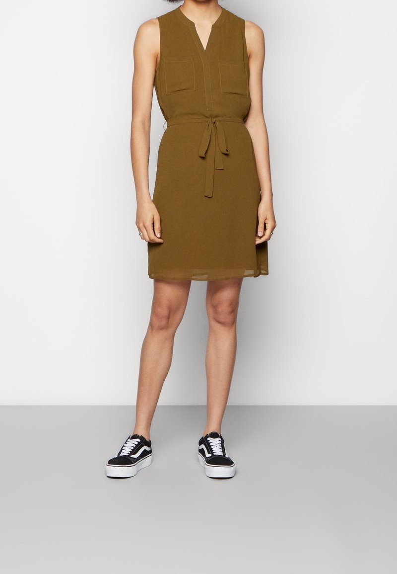 Robe olive sans manches avec poches avant et ceinture à la taille, portée avec des baskets plateformes noires et blanches sur une personne debout.