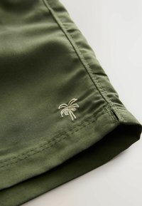 Rohelised riidest püksid, millel on all ääres väike beež tikitud palmi logo, sileda, mati tekstuuri ja peene õmblusega.