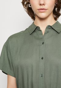 Camicetta verde oliva a maniche corte con colletto, dotata di bottoni e texture del tessuto liscia, mostrata fino al collo.