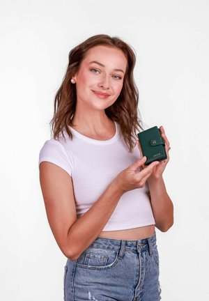 Zelená kožená peněženka s uzávěrem na knoflík, obsahující značkový akcent. Modelka předvádí peněženku, obléká bílé přiléhavé tričko a džínové šortky.