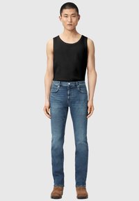 Man standing wearing a black sleeveless shirt, blue slim-fit jeans, and brown suede boots on a plain light gray background.