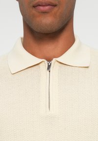 Maglione color crema con lavorazione a maglia testurizzata, dotato di un colletto con zip e un colletto corto a stola. Il primo piano mette in evidenza i dettagli del colletto.