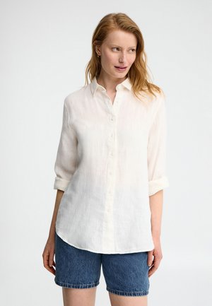 Femme portant une chemise blanche à manches longues avec les poignets retroussés et un short en denim bleu, debout devant un fond clair uni.