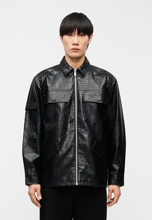 Mann trägt eine schwarze Jacke aus Kunst-Krokodilleder mit Frontreißverschluss und vier Klappentaschen, steht vor einem schlichten hellen Hintergrund.