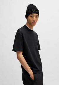Svart t-skjorte med korte ermer, minimalistisk design og lomme, sammen med en ribbet svart lue med ordet "HUGO."