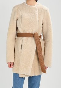 Abrigo de piel sintética beige con cinturón en la cintura, que presenta un cuello redondo, mangas largas y dos bolsillos delanteros. Detalle de cinturón de cuero liso.