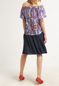 Blusa colorata senza spalle con motivi floreali e geometrici, abbinata a una gonna plissettata navy e scarpe ballerine rosse. Tessuti morbidi.
