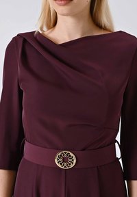 Abito bordeaux con maniche a tre quarti, caratterizzato da un scollo drappeggiato e una fibbia circolare dorata con un design intricato. Tessuto dalla superficie liscia.
