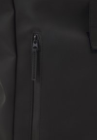 Detail černého voděodolného batohu s uzávěrem na zip, hladkým matným povrchem a zesíleným prošíváním kolem oblasti zipu.