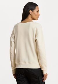 Una donna con lunghi capelli scuri indossa una felpa color crema e pantaloni neri, vista da dietro e con sguardo rivolto a destra.