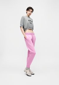 Νεαρός/ή στέκεται με τα χέρια στις τσέπες, φορώντας γκρι cropped φούτερ Nike, ροζ φόρμα, γκρι κάλτσες και λευκά αθλητικά παπούτσια, με απλό φόντο.