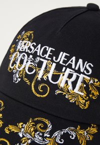 Black cap featuring ornate gold and white embroidery, displaying the text "VERSACE JEANS COUTURE" with decorative patterns.
