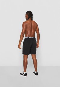 Pantaloni scurți de baie negri cu un brâu elastic alb pe care este inscripționat textul "URBASICS", asortați cu adidași negri și un fundal simplu.