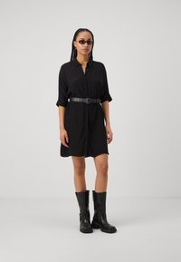 Schwarzes Hemdkleid mit durchgehender Knopfleiste, aufgerollten Ärmeln, gerafft mit einem schwarzen Nietengürtel, kombiniert mit schwarzen kniehohen Stiefeln.
