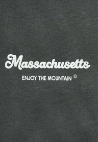 Tessuto grigio con testo bianco. Nella parte superiore è riportato "Massachusetts" in un font stilizzato, e sotto si legge "ENJOY THE MOUNTAIN ©."