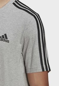 Camiseta de algodón gris con logo negro de Adidas, cuello redondeado y tres franjas negras en cada hombro. Mangas cortas.