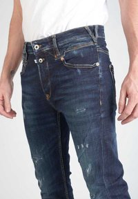 Jeans de denim azul oscuro con un ajuste slim, acentos desgastados y costuras de contraste en las costuras, con cinco bolsillos y cierre de botón.