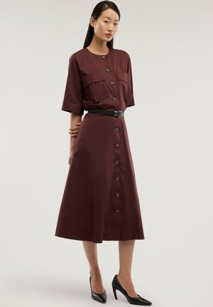 Frau trägt ein braunes Midikleid mit kurzen Ärmeln und Knöpfen, einen schwarzen Gürtel sowie schwarze High Heels und steht vor einem schlichten Hintergrund.