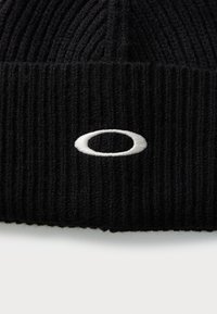 Schwarze, gestrickte Mütze mit vertikalen Rippen. Besitzt ein weißes gesticktes Logo auf der Vorderseite. Das Material wirkt weich und strukturiert.