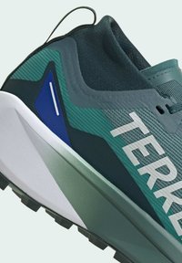 Scarpa da ginnastica verde con tomaia testurizzata, accento blu e logo bianco. La suola esterna presenta tasselli di trazione per stabilità.