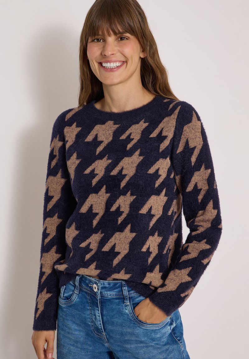 Femme souriante aux cheveux bruns portant un pull bleu marine avec un grand motif pied-de-poule beige et un jean bleu, debout devant un fond uni.