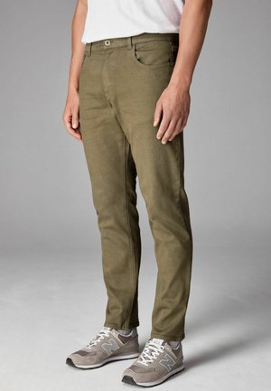 Pantalons fuselés vert olive en tissu doux, dotés d'un design classique à cinq poches et d'une fermeture à bouton à l'avant, associés à des baskets grises.
